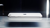 A modern smartphone rests horizontally, showcasing a sleek and minimalist design with a metallic frame and smooth edges. The camera lens is visible, and the iconic brand logo is centered. The lighting emphasizes the phone's polished surface.